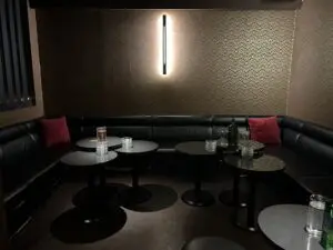 Dimly lit lounge area with a black curved sofa, small round tables, red cushions, water bottles and glasses, and a vertical wall light.