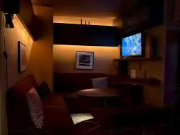 Cozy, dimly lit lounge with a round table, red sofa and patterned cushions, framed photo on the wall, and warm wall lights.