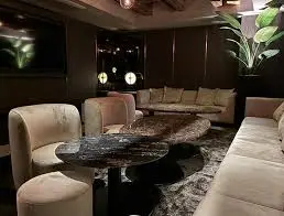 Dim hotel lounge with curved beige sofas, round black marble tables, and a large plant in the corner.