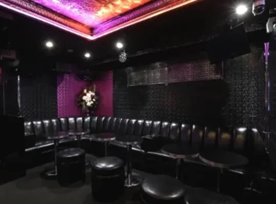 Dimly lit club lounge with curved black leather seating, round stools, small tables, purple ceiling lighting, and a flower arrangement on a purple wall.