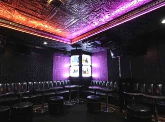 Club lounge with black circular stools and curved seating, purple lighting, and a central display screen at a dimly lit venue.