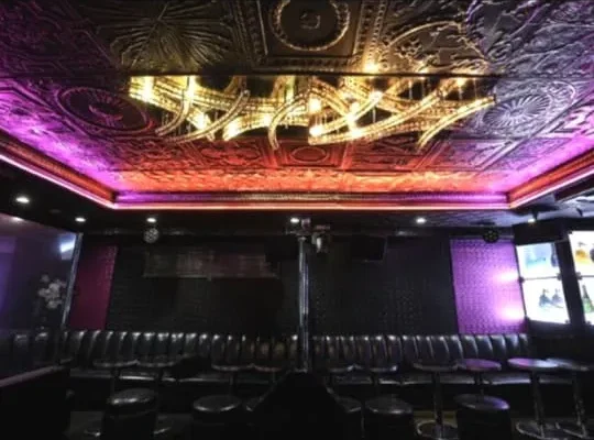 Dim nightclub lounge with an ornate ceiling, purple uplighting, long black leather booths, and small round tables.