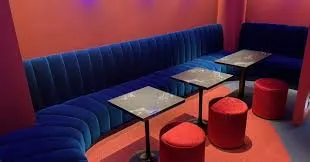 Blue curved upholstered booth along a red wall with small square tables and red round stools, forming a cozy lounge seating area.