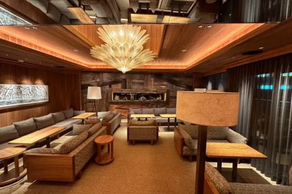 Luxurious hotel lounge with wood-paneled ceiling, a large feather chandelier, and L-shaped sofas around wooden tables.] ,