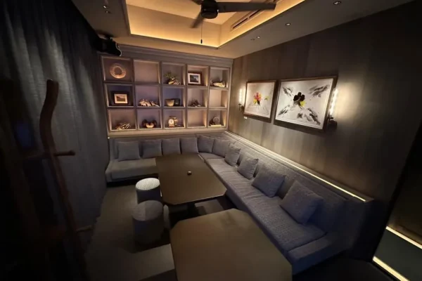Dimly lit lounge with an L-shaped gray sofa around two brown coffee tables and a back wall of decorative shelves.
